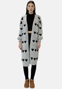 Długi szary kardigan z czarnymi wzorami serc, o dzianinowej fakturze, z otwartym przodem i ściągaczami na rękawach, noszony na czarnym crop topie i legginsach.
