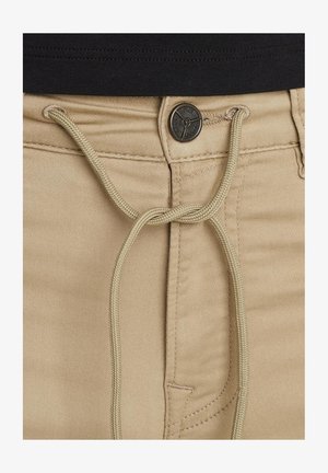 Beige katoenen broek met een trekkoordsluiting, ritssluiting en een ronde zwarte metalen knop met reliëfdesign details.