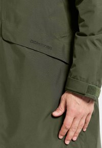 Olijfgroene waterdichte jas met een gestructureerd oppervlak, zichtbare stiksels en een klittenbandsluiting aan de manchetten. Merkennaam zichtbaar op de stof.