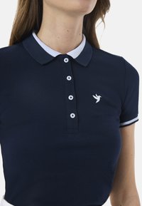 Polo di colore blu navy con colletto e bordi bianchi, dotato di cinque bottoni bianchi e di un piccolo logo a forma di uccello bianco sul lato sinistro del petto. Tessuto testurizzato.