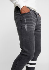 Dark gray denim jeans with distressed details, visible stitching, and a tapered fit. Features white striped accents near the lower leg.