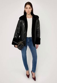 Černá kožená bunda s kožešinovým límcem, sladěná s úzkými modrými džínami a vínovými lodičkami, držící malou černou kabelku.
