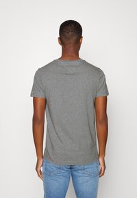 Man med kort lockigt hår som bär en enkel grå kortärmad t-shirt och ljusblå jeans, står vänd mot en vit vägg.