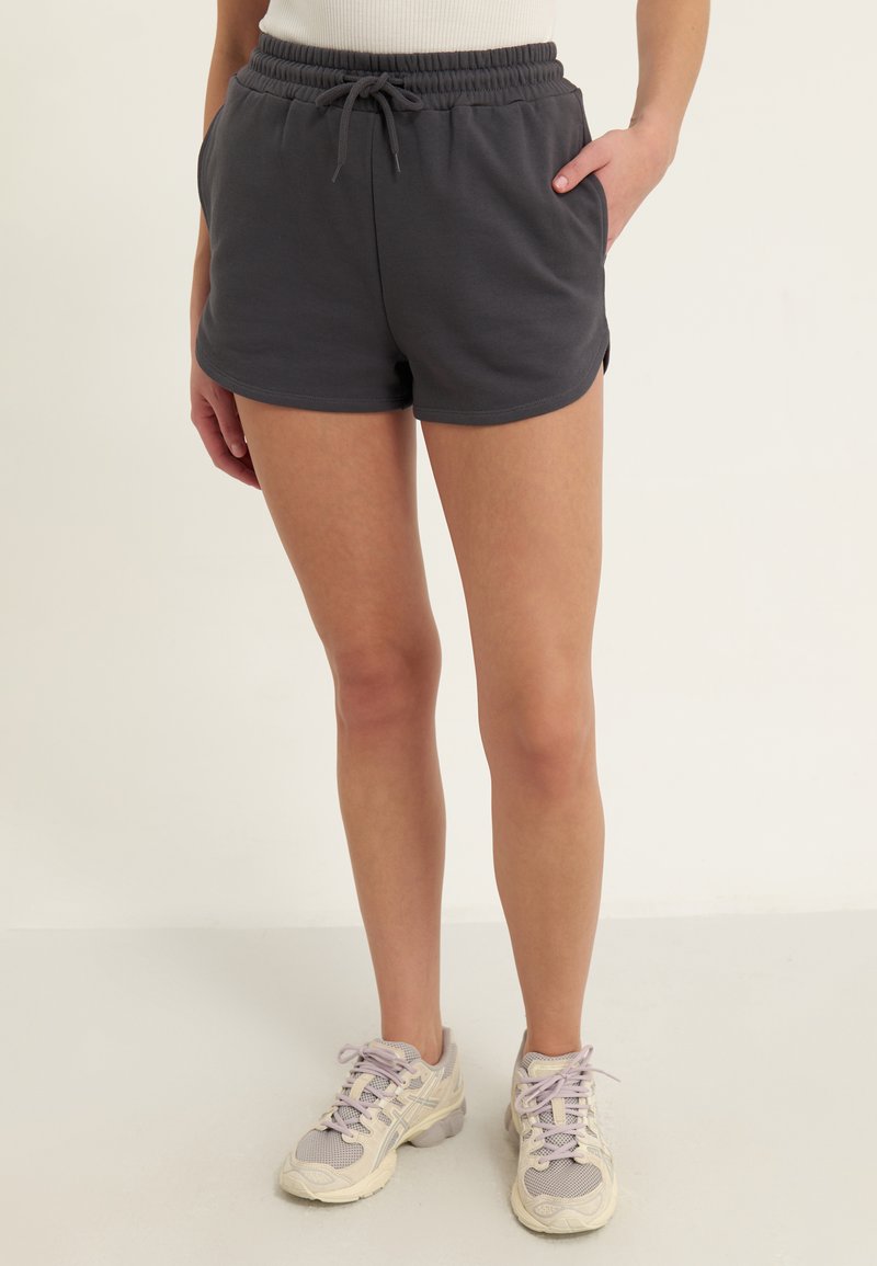 Personne portant un short élastique gris foncé à la taille et des chaussures de sport lacées de couleur claire, debout sur un fond clair.
