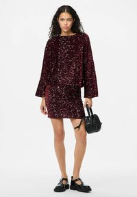 Top burdeos con lentejuelas y mini falda a juego con mangas sueltas. La modelo sostiene un bolso circular negro y lleva unos zapatos de plataforma negros.