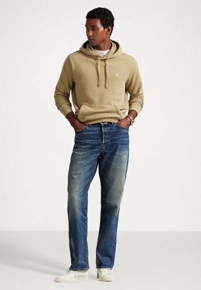 Man in een beige hoodie, vervaagde blauwe jeans, witte sneakers en een horloge, staand met zijn handen in de zakken en kijkend naar de zijkant.