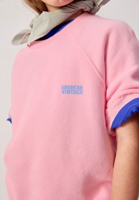 Personne portant un sweat-shirt rose avec un logo bleu « AMERICAN VINTAGE », des poignets de manches bleus et une écharpe à carreaux grise autour du cou.