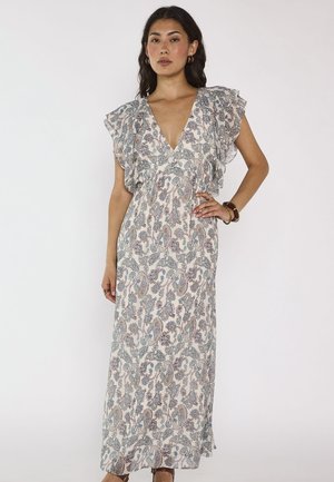 Femme portant une longue robe sans manches à col en V, épaules à volants et imprimé cachemire aux tons pastels, debout devant un fond blanc uni.