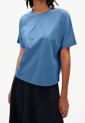 Person trägt ein blaues T-Shirt mit kurzen Ärmeln und Rundhalsausschnitt und einen schwarzen Rock vor einem schlichten weißen Hintergrund.