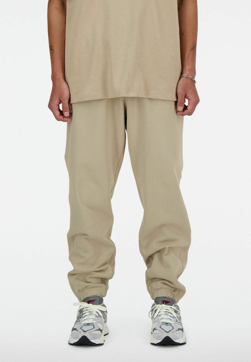 Partie inférieure d'une personne portant un pantalon de survêtement beige avec des poignets élastiques et des baskets grises, blanches et rouges, se tenant devant un fond blanc uni.