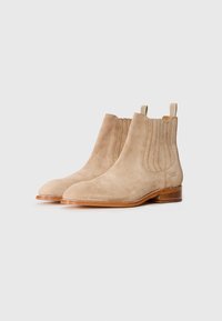 Botas Chelsea de ante beige con detalles acanalados y puntera curvada. Tacón plano de madera y pequeña tirador en la parte posterior.