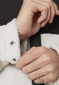 Chaqueta de forro polar blanca con detalles negros, que presenta un cierre a presión y una tela texturizada en los puños, mostrando manos ajustando la chaqueta.