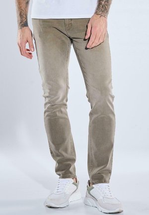 Man wearing fitted beige pants, white sneakers, and a white shirt, standing against a plain light background.