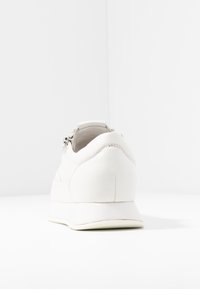 Valkoinen nahkainen lenkkarit, sileä pinta. Ominaisuuksiin kuuluu pyöreä kärki, hillitty ompele, ja pieni korko. Takana vetoketju helpottaa pukemista.