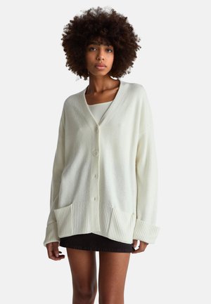 Cremefarbener Cardigan mit V-Ausschnitt, Knopfleiste, geripptem Saum und Bündchen sowie zwei Fronttaschen. Weiche Textur und oversized Passform.