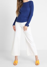 Niebieski prążkowany sweter z długimi rękawami i guzikami, zestawiony z szerokimi białymi spodniami i żółtymi sandałami na otwartym palcu.