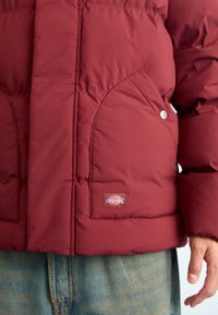 Maroon puffer jacket with a quilted design, featuring a side pocket and a branded patch. Smooth fabric with a glossy finish.