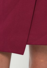 Φούστα τύπου wrap σε μπ ορντό με υφή υφάσματος. Διαθέτει ασύμμετρο σχέδιο και καθαρές γραμμές, προσφέροντας μια ομαλή, ραμμένη εμφάνιση.