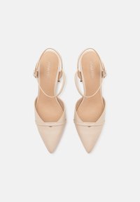 Dei tacchi slingback in pelle verniciata beige con punta appuntita, cinturino alla caviglia e dettagli di cucitura minimi sulla parte superiore.