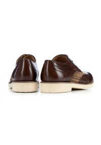 Bruine leren schoenen met een gladde afwerking, met patroonaccenten aan de zijkant en een romig-witte rubberen zool, gezien vanaf de achterkant.