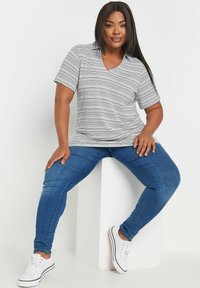 Grå och vit randig kortärmad skjorta med V-ringning, i kombination med blå skinny jeans och vita sneakers, sittande på en vit plattform.