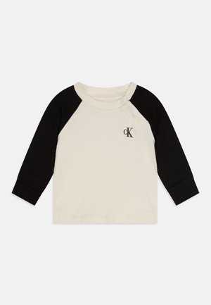 Camisa de algodão de manga longa com corpo em creme e mangas raglan pretas. Apresenta um botão no ombro e um pequeno logótipo no peito.