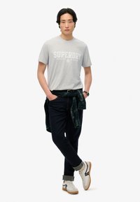 Grå t-shirt med "SUPERDRY" logo, parret med mørk denim jeans og hvide sneakers. En grøn plaid skjorte er bundet omkring taljen.