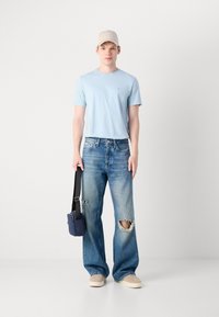 Hellblaues T-Shirt, ausgebeulte weite Jeans in verblasstem Blau, beigen Sneakers und einer beigen Cap. Schwarze Schultertasche aus Textil abgebildet.