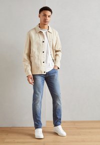 Light beige jacket with a button front, white t-shirt underneath, blue jeans, and white sneakers standing on a wooden floor against a light gray wall.