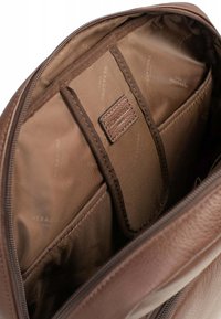 Wnętrze brązowej skórzanej torby z gładką, ciemniejszą podszewką, wieloma kieszeniami oraz małą skórzaną metką marki.