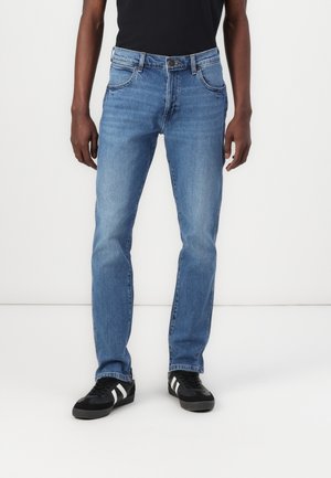 Man wearing light blue straight-fit jeans and black sneakers with white stripes, standing against a plain white background.