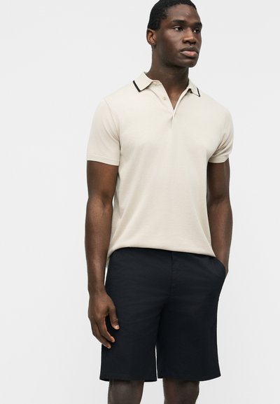 Homme portant un polo beige à manches courtes avec des bordures noires et un short noir au genou, debout devant un fond uni.