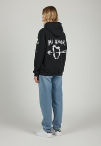 Felpa nera con grafica bianca "Mi Amor" e frecce sulla schiena, abbinata a jeans azzurri chiari e sneakers bianche con dettagli neri.