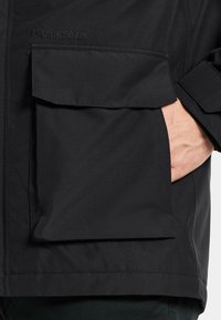 Svart vattentät jacka med en stor framficka, slät textur, minimalistisk design och detaljerad varumärkesnamn "Didriksons" på tyget.