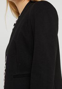 Musta bleiseri, jossa on jämäkät olkapäät, rintatasku ja teksturoidusta kankaasta valmistettu pinta. Sivukuva korostaa räätälöityä istuvuutta ja suunnittelun yksityiskohtia.