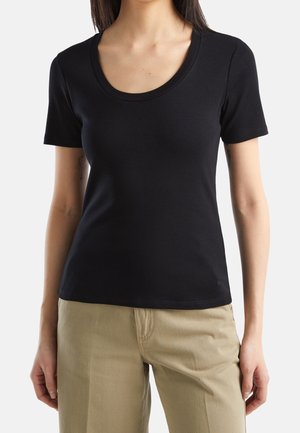 Femme portant un t-shirt noir moulant à manches courtes et col rond, ainsi qu’un pantalon beige, vue des épaules jusqu’aux hanches sur un fond uni.
