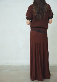 Personne avec de longs cheveux bruns portant un pull en tricot marron à manches courtes et une jupe plissée longue assortie, se tenant contre un mur clair.