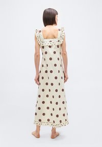 Γυναίκα που στέκε ται ξυπόλυτη και γυρίζει την πλάτη, φορώντας ένα κρεμ αμάνικο φόρεμα με μεγάλα καφέ πουά και βολάν στη λαιμόκοψη και στο τελείωμα.