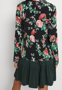Mujer vistiendo un blazer negro ajustado con estampado floral en rojo y rosa y una falda plisada verde oscuro, mostrada desde atrás.