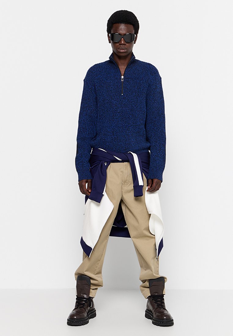 Blauer Strickpullover mit halbem Reißverschluss, beigefarbene Cargo-Hosen mit weißen Akzenten, dunkle Sonnenbrille und schwarze klobige Stiefel. Lässiger, geschichteter Look.