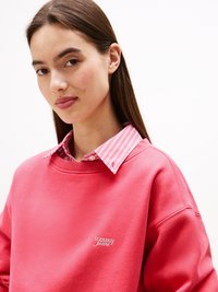Felpa rosa con scollo rotondo e logo ricamato "tommy jeans", indossata sopra una camicia a righe nei colori rosa e bianco.