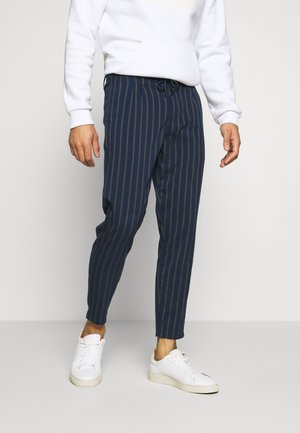 Mann trägt marineblaue Nadelstreifenhose, weißen Hoodie und weiße Turnschuhe, steht vor einem einfarbigen hellen Hintergrund.