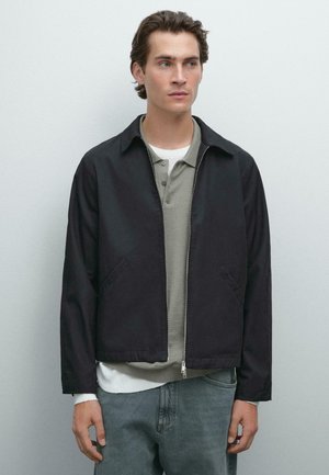 Dark navy zip-up jacket with a structured collar, two side pockets, and a smooth texture, layered over a light gray polo and white shirt.