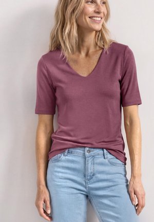 Femme souriante portant un t-shirt mauve à manches courtes et col en V et un jean bleu clair sur un fond neutre.