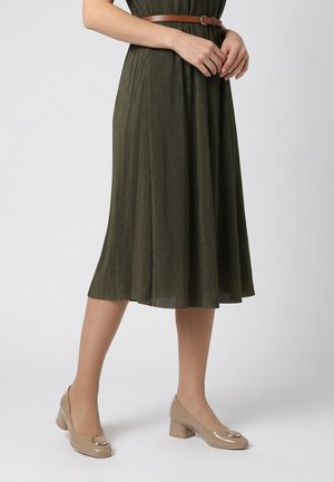 Vrouw draagt een olijfgroene geplooide midi-rok met een bruine riem en beige lakleren enkellaarsjes met blokhak en metalen details.