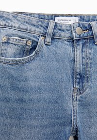 Jean en denim bleu clair avec poche avant, passants pour ceinture, bouton en métal et étiquette de marque visible à l'intérieur de la ceinture.