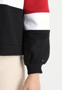 Pullover nero, rosso, bianco e giallo con colletto e polsini a coste. La manica presenta un dettaglio arricciato al polso. Logo visibile.