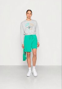 Sweatshirt cinza com impressão "LON YORK", combinado com calções verdes, ambos feitos de tecido macio. O modelo usa ténis brancos e segura uma bolsa verde.