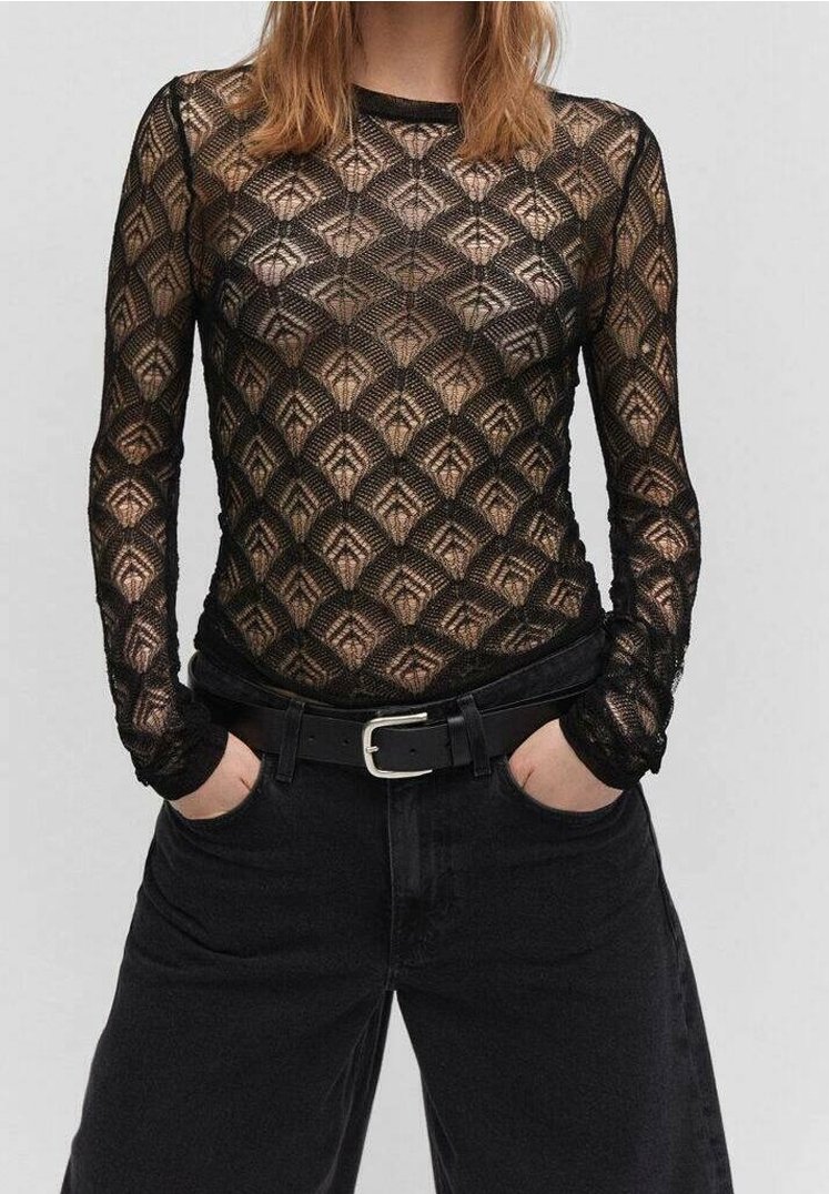 Femme portant un haut en dentelle noire à manches longues avec des motifs géométriques, un pantalon noir taille haute et une ceinture noire avec une boucle argentée.
