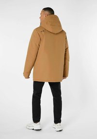 Braune gepolsterte Jacke mit Kapuze, die eine glatte Textur und ein Logodetail auf dem Rücken hat. Kombiniert mit schwarzen Jeans und weißen Sneakern.
